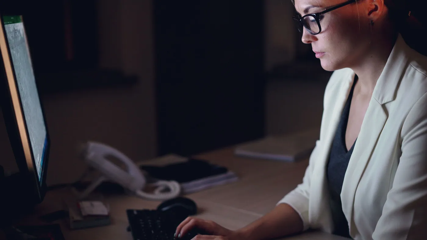 A woman in glasses working late at a desk lit only by her computer monitor