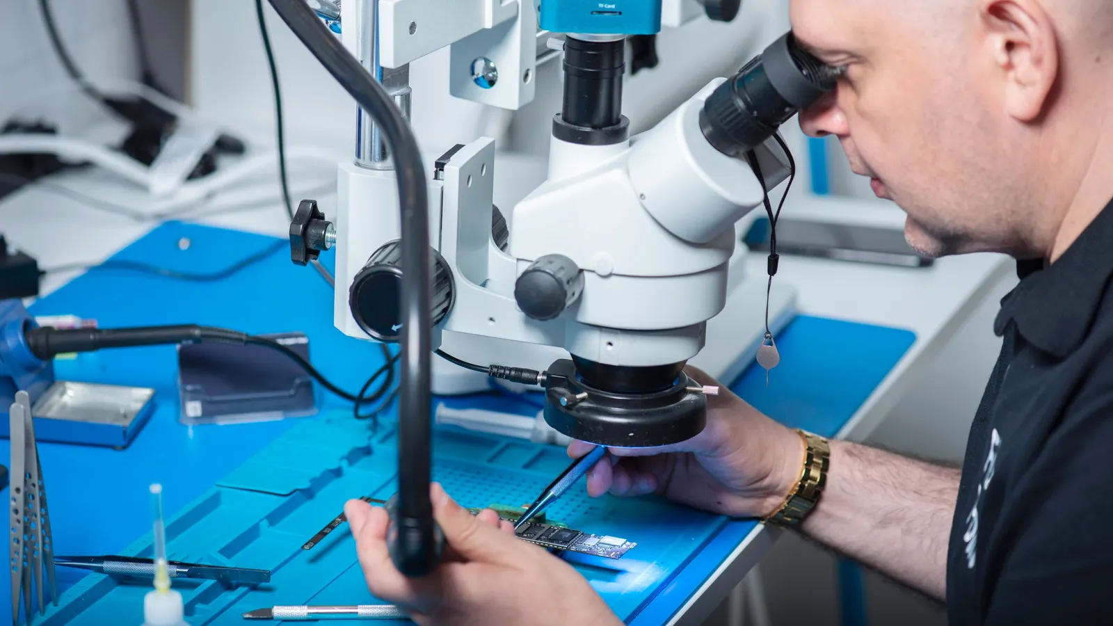 A technician looks through a stereo microscope while holding a circuit board under a ring light