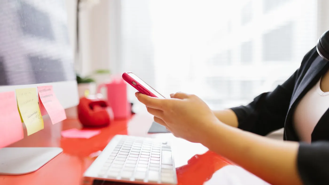 A person at an office desk with sticky notes checking a message on a red smartphone