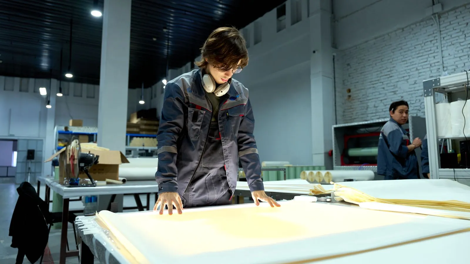 A worker in uniform inspecting sheets under bright light in a factory