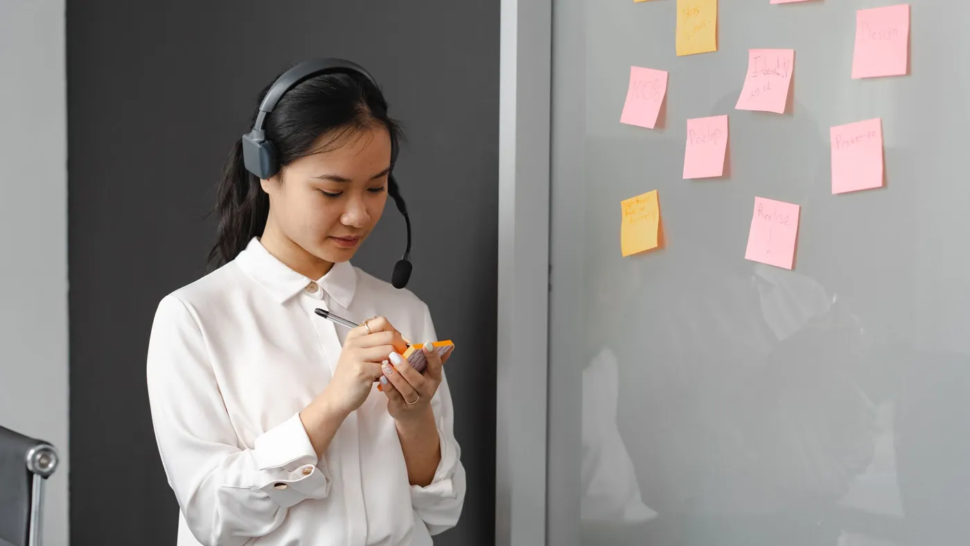 A support agent in a headset writing on a pad of sticky notes beside a glass wall covered in more notes