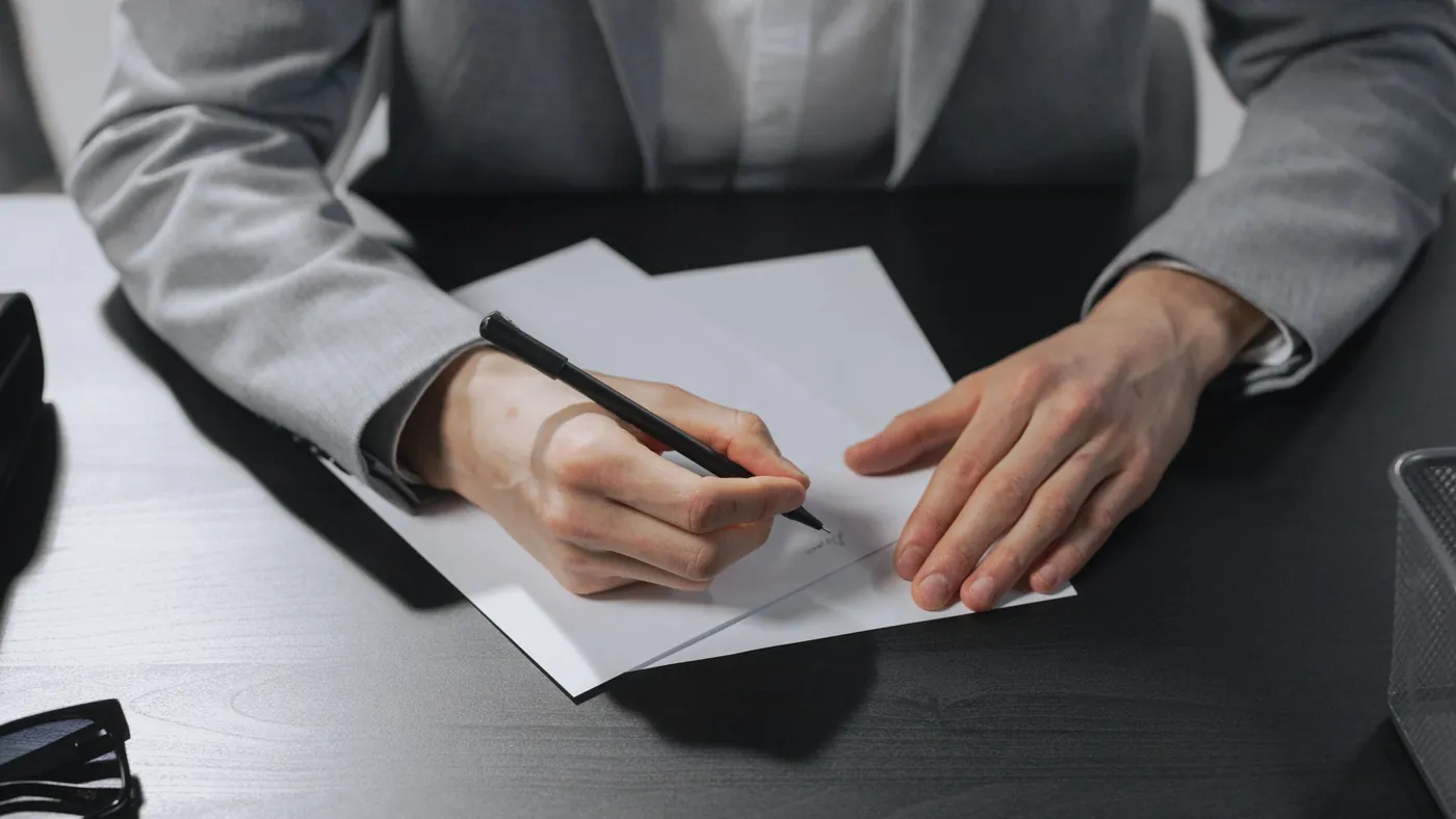 A person in a grey suit signing a sheet of paper at a dark desk
